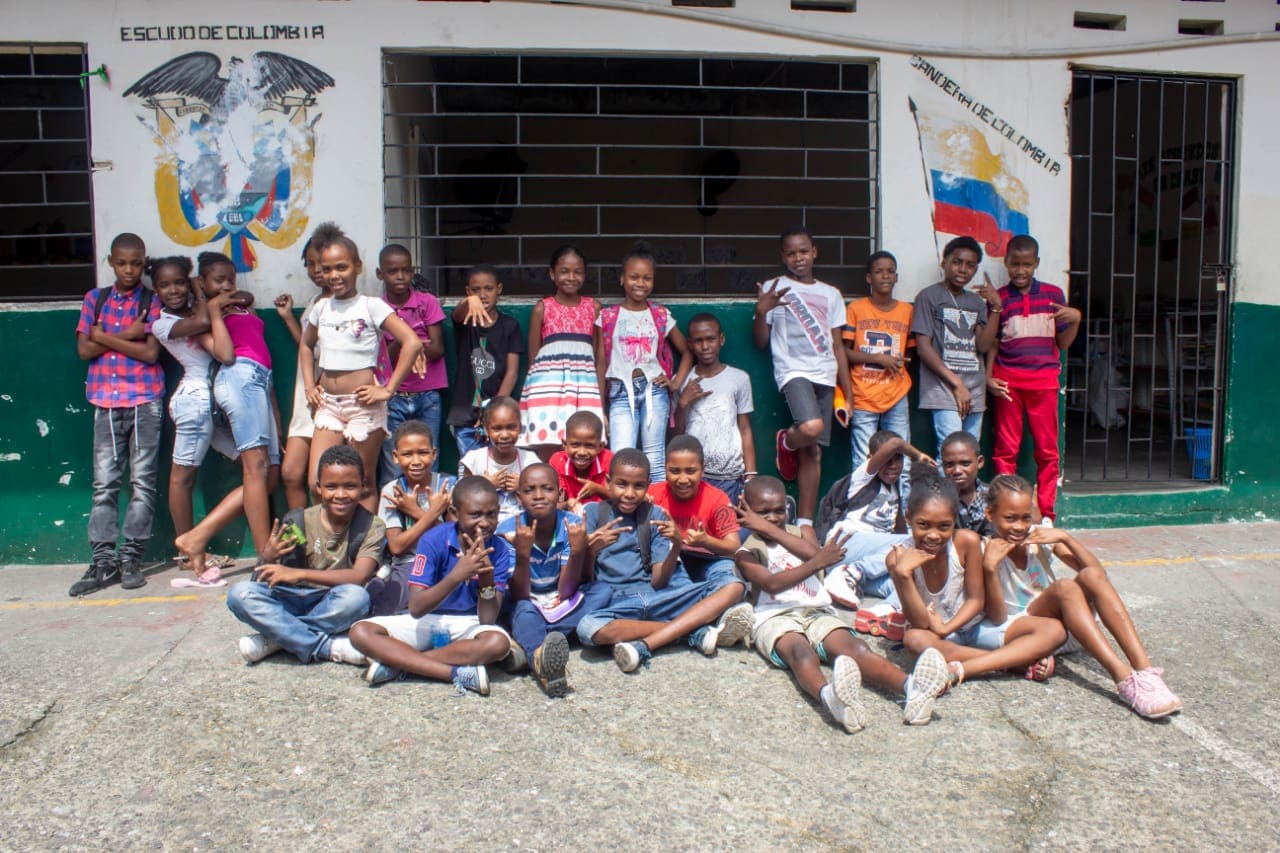la imagen puede contener a un grupo de niños,algunos sentados en el piso y otros de pie en la parte de atras.